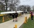 Rowers transport a boat along a path, surrounded by trees and spectators at a rowing event.