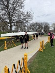 Rowers transport a boat along a path, surrounded by trees and spectators at a rowing event.