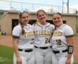 Three female softball players in team uniforms smile together on the field, showcasing team spirit and camaraderie.