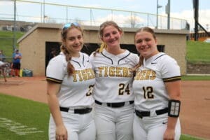 Three female softball players in team uniforms smile together on the field, showcasing team spirit and camaraderie.
