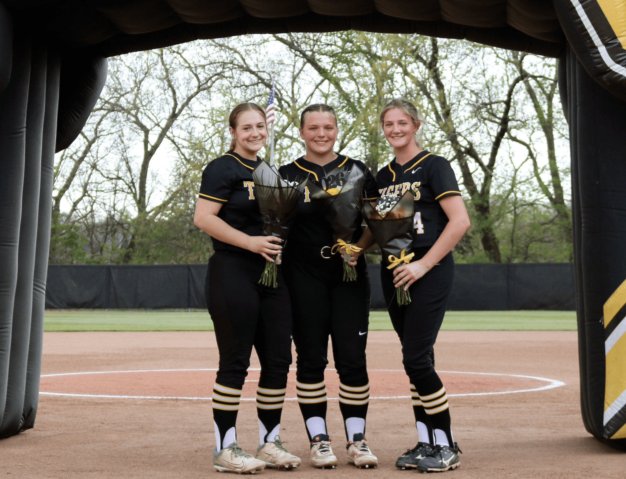 Three softball players pose at the field's entrance, each holding a bouquet, celebrating their achievements together.