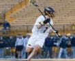 Lacrosse player wearing white jersey and helmet, executing a shot on goal during a game. Stadium spectators in background.