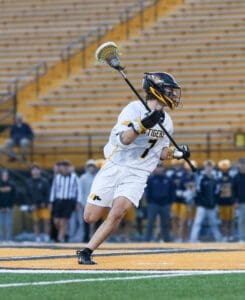 Lacrosse player wearing white jersey and helmet, executing a shot on goal during a game. Stadium spectators in background.