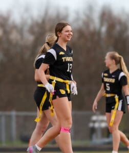 Cheerful female athlete in a flag football uniform, smiling on the field with teammates during practice.