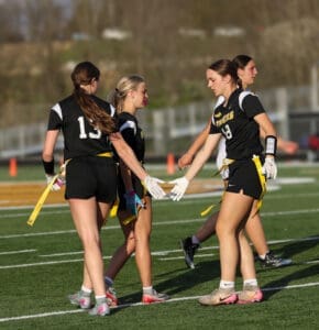 Female athletes in black uniforms strategizing during a flag football game on a sunny field. Teamwork in action!