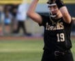 A softball player in a black uniform prepares to pitch, holding the ball and glove above her head on the field.