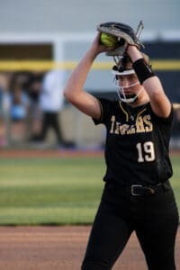 A softball player in a black uniform prepares to pitch, holding the ball and glove above her head on the field.