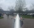 Two rowers hold a boat, preparing for practice on a cloudy day at a rowing event near parked cars and a grassy area.