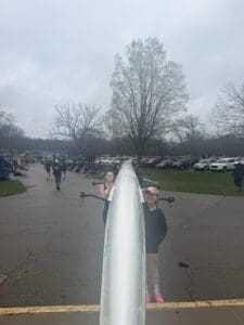 Two rowers hold a boat, preparing for practice on a cloudy day at a rowing event near parked cars and a grassy area.