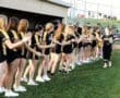 Team members in black and gold uniforms celebrate and greet players before a game on a grassy field.