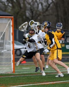 Lacrosse players in action as one goalie attempts to block a shot while teammates prepare for the play.