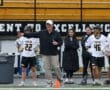 Lacrosse coach instructs players on the field, with team members in uniforms and spectators in the background.