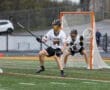 Lacrosse players in team uniforms compete on the field, with one player preparing to shoot on goal.