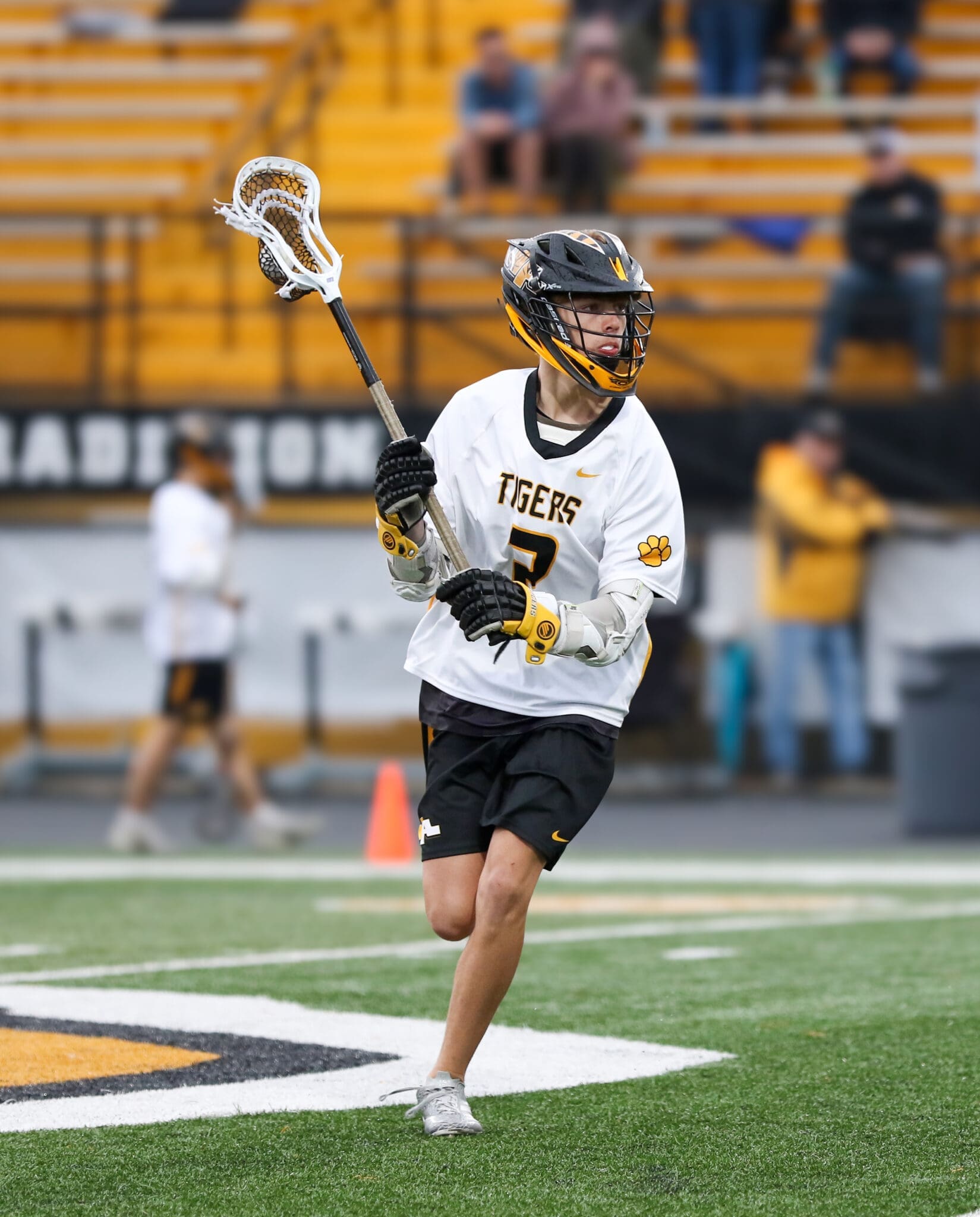 Lacrosse player in action wearing a Tigers jersey, dribbling on the field with spectators in the background.
