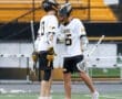 Two young lacrosse players celebrate on the field, showcasing teamwork in their white and yellow uniforms.
