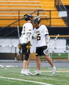Two young lacrosse players celebrate on the field, showcasing teamwork in their white and yellow uniforms.
