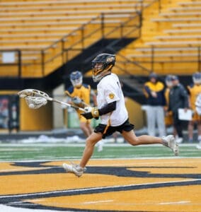 Young lacrosse player in action during a practice session on a field with yellow and black markings, teammates in background.