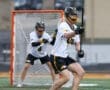Lacrosse players in action on a field, one preparing to shoot while the goalie guards the net.