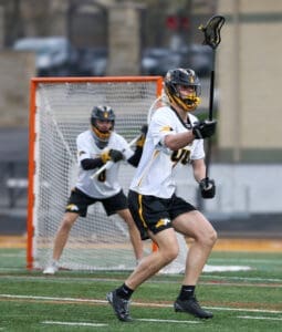 Lacrosse players in action on a field, one preparing to shoot while the goalie guards the net.