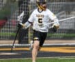 Lacrosse player in action, wearing a Tigers jersey, sprinting on field with a stick and helmet during a game.