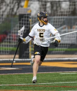 Lacrosse player in action, wearing a Tigers jersey, sprinting on field with a stick and helmet during a game.