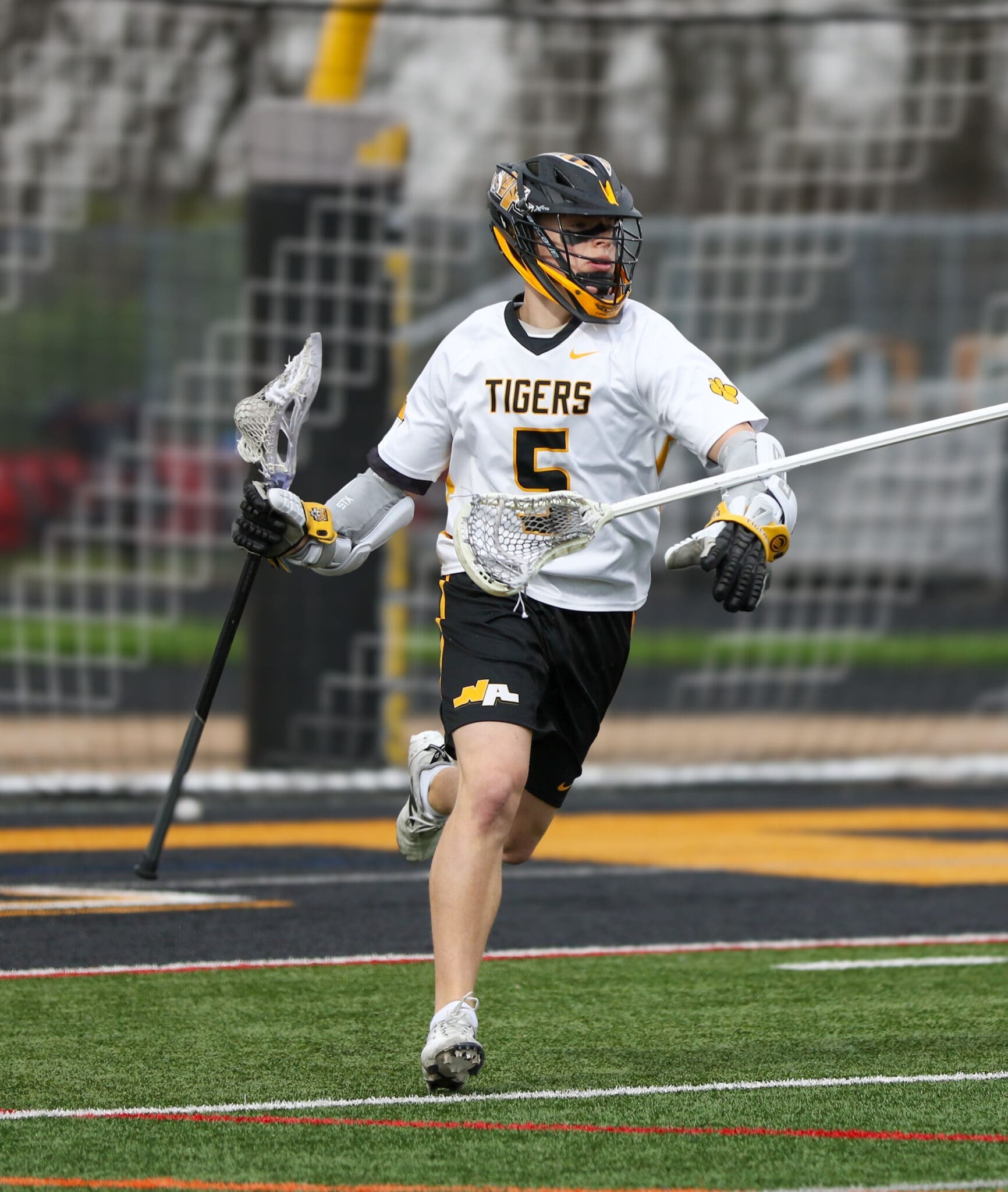 Lacrosse player in action, wearing a Tigers jersey, sprinting on field with a stick and helmet during a game.