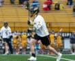 Lacrosse player running with stick on field during a game, while spectators in the background hold umbrellas.