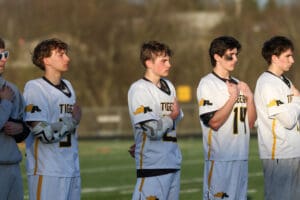 Lacrosse players in jerseys stand attentively during the national anthem on the field. Team spirit and unity on display.