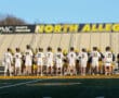 Lacrosse players in uniforms gather on the field at North Allegheny High School as a referee oversees the match.