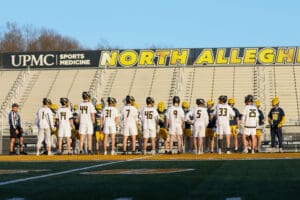 Lacrosse players in uniforms gather on the field at North Allegheny High School as a referee oversees the match.