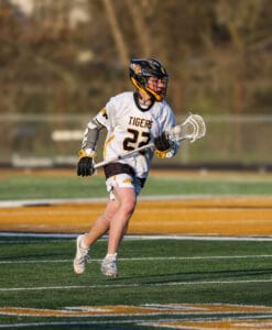 Lacrosse player wearing a Tigers jersey runs on the field, holding a stick during a competitive match.