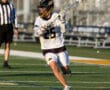 Lacrosse player in white uniform #25 prepares to pass the ball on a sunlit field, with an official observing nearby.