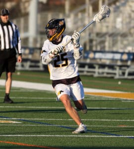 Lacrosse player in white uniform #25 prepares to pass the ball on a sunlit field, with an official observing nearby.