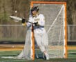 Lacrosse goalie in action, wearing team jersey and gear, preparing to block a shot at the goal.