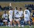 Lacrosse players in team jerseys celebrate a goal, with fans cheering in the background at a lively sports event.