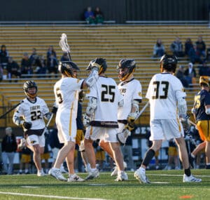 Lacrosse players in team jerseys celebrate a goal, with fans cheering in the background at a lively sports event.