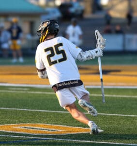 Lacrosse player wearing jersey number 25, sprinting on a field with Brotherhood on his shorts, focused on the game.
