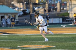 Lacrosse player in action on the field, wearing a white jersey, running with a stick, showcasing athleticism and teamwork.