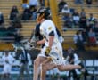 High school lacrosse player in white uniform sprinting with a lacrosse stick, surrounded by spectators in the stands.