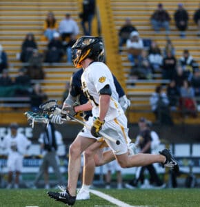 High school lacrosse player in white uniform sprinting with a lacrosse stick, surrounded by spectators in the stands.