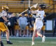 Player in white lacrosse uniform performs a shot while another player defends, showcasing action on the field.