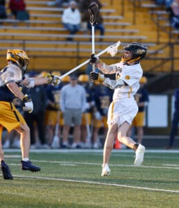 Player in white lacrosse uniform performs a shot while another player defends, showcasing action on the field.
