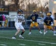 Lacrosse players compete on a field; one player prepares to pass while defenders position themselves nearby.