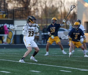 Lacrosse players compete on a field; one player prepares to pass while defenders position themselves nearby.