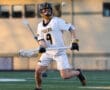 Lacrosse player in action, wearing a jersey and helmet, preparing to catch a ball on the field during a game.