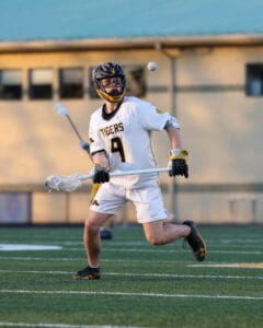 Lacrosse player in action, wearing a jersey and helmet, preparing to catch a ball on the field during a game.
