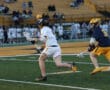 High school lacrosse players compete on the field, one dodging an opponent while holding a lacrosse stick.