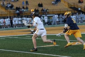 High school lacrosse players compete on the field, one dodging an opponent while holding a lacrosse stick.