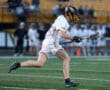 Lacrosse player sprinting on the field, stick in hand, wearing a helmet and team uniform during a game.