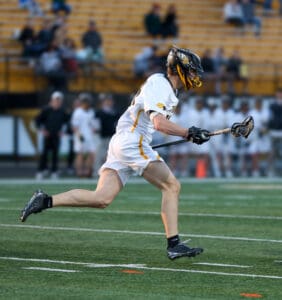 Lacrosse player sprinting on the field, stick in hand, wearing a helmet and team uniform during a game.